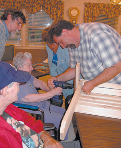 Resident Jean Fortney helps with chair assembly. Her son, Dan Fortney watches as volunteers Carol Briggs and Larry Frazee help guide the drill.