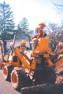 Rod Mayo, top and Nick Fisher operate a combination trencher and backhoe at 2427 Allegany Avenue on March 24, 2005.  The trencher digs the trench, the four-inch orange conduit is placed in the ground and the backhoe covers it up.  Two men, on hand shovels, do the clean-up work and later another crew will pull up to three black fiber-optics cables through the four-inch trace.