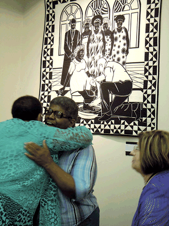 Quilter’s Hall of Fame inductee Carolyn Mazloomi receives a hug from a well-wisher as they stand in front of her quilt In the spirit of forgiveness.